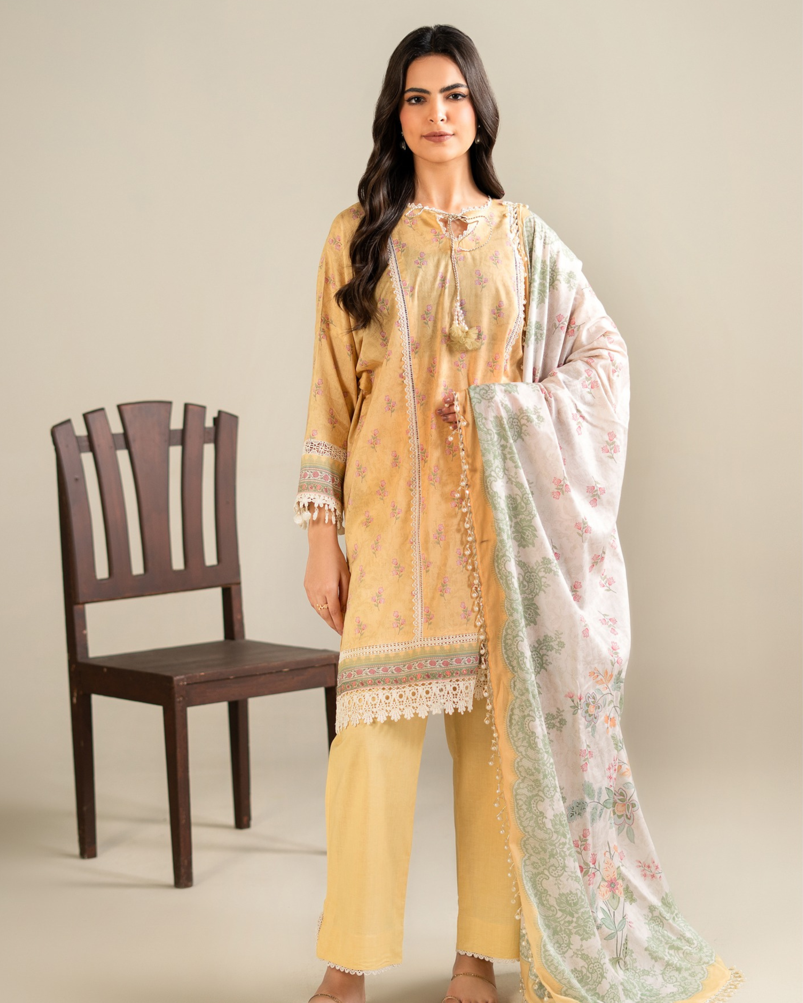 Woman wearing a yellow traditional outfit with a floral dupatta, standing next to a wooden chair.