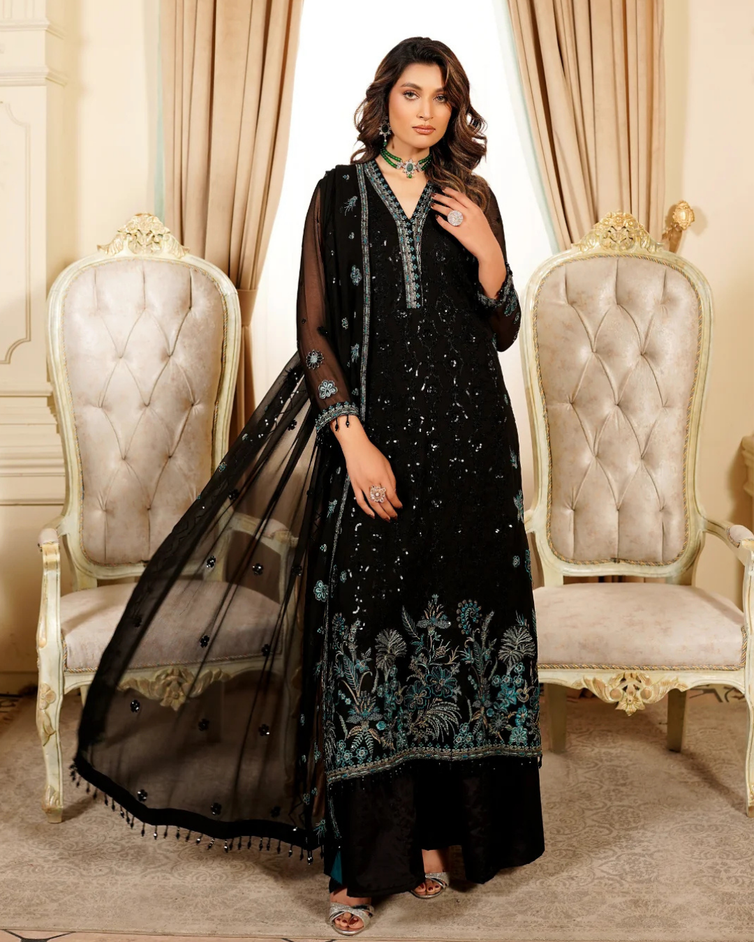 Woman in a black embroidered outfit standing between two ornate chairs.