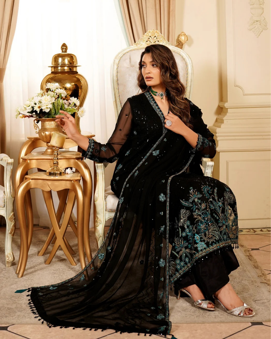 Woman in a black embroidered saree sitting in an elegant room with gold decor.