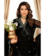 Woman in a black embroidered saree holding a vase with flowers, wearing a green necklace.