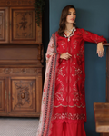 Woman in a red embroidered traditional outfit standing against a blue wall with wooden furniture.