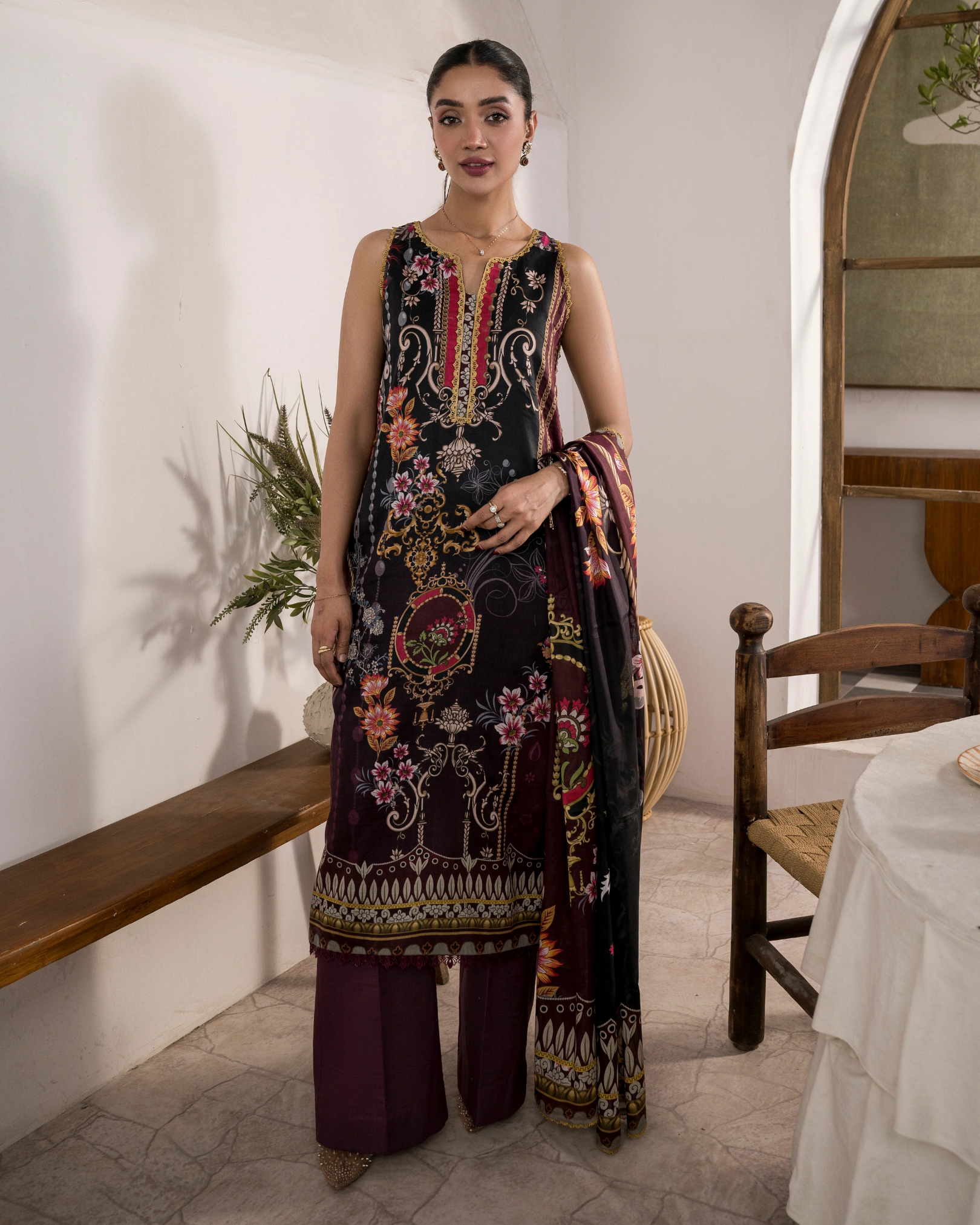 Woman in an embroidered outfit standing in a room with wooden furniture and a plant.