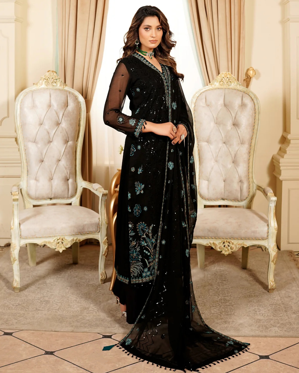 Woman in a black embroidered saree standing in an elegant room with white chairs and beige walls.