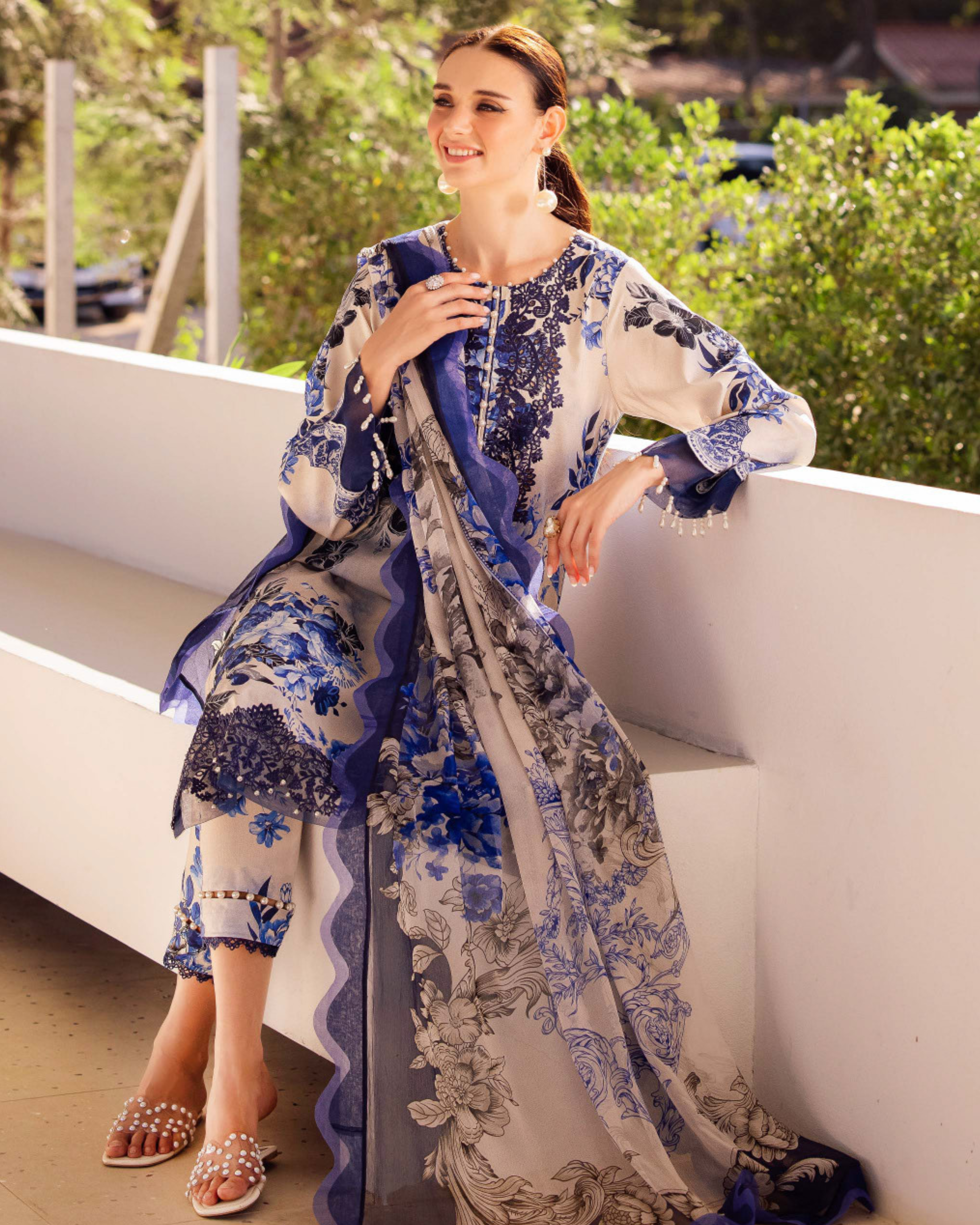 Woman in a blue and white floral outfit sitting on a white ledge with greenery in the background
