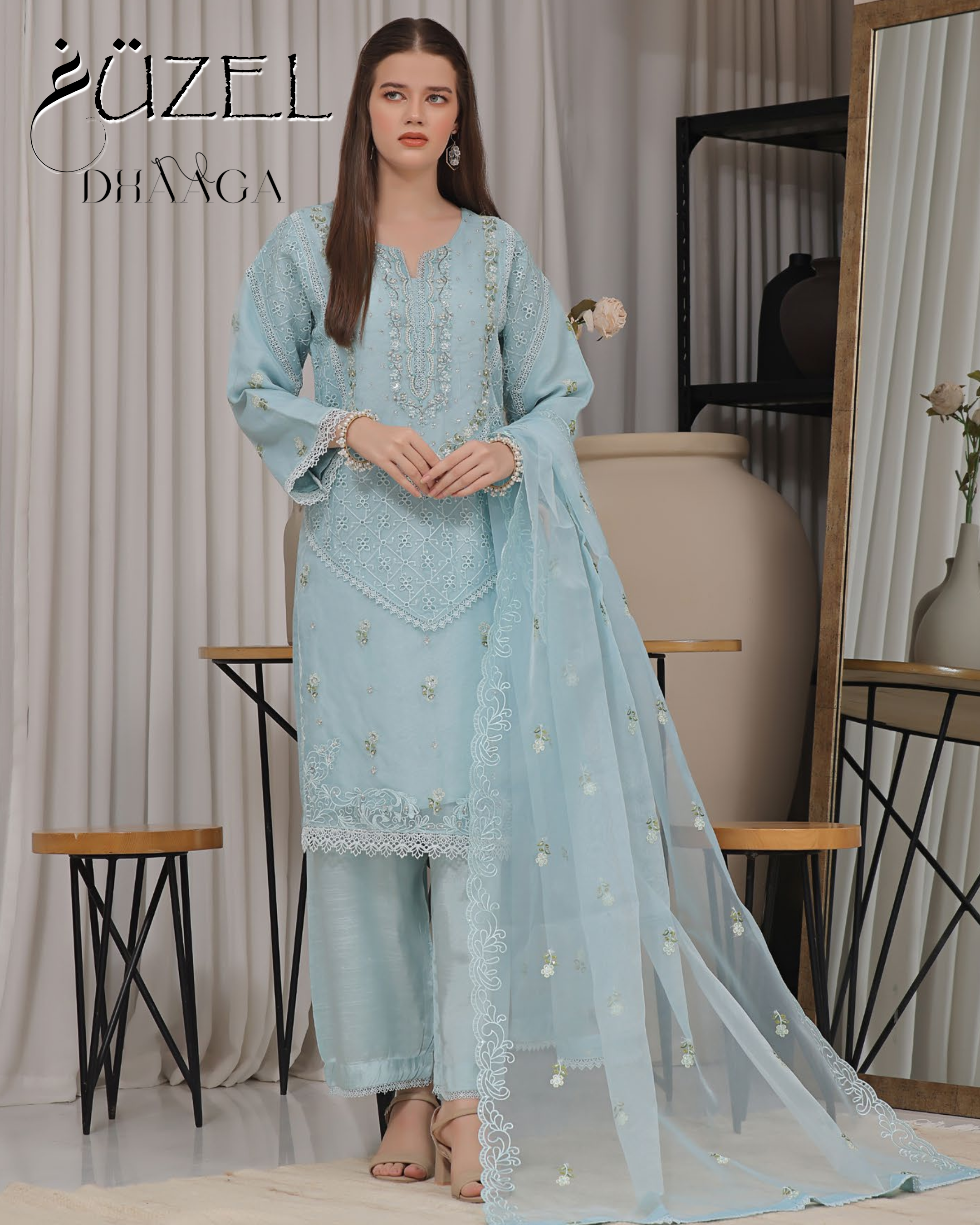 Woman wearing a light blue embroidered outfit in a room with a mirror and furniture.