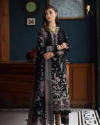 Woman wearing a black embroidered dress in an indoor setting with wooden furniture and decorative elements.