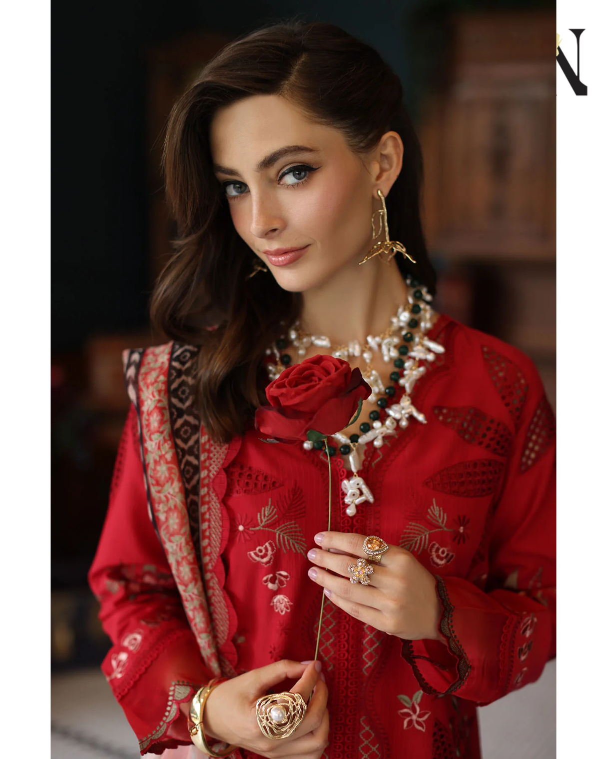 Woman in a red embroidered outfit holding a rose, wearing jewelry.