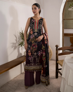 Woman in an embroidered outfit standing in a room with wooden furniture and a plant.