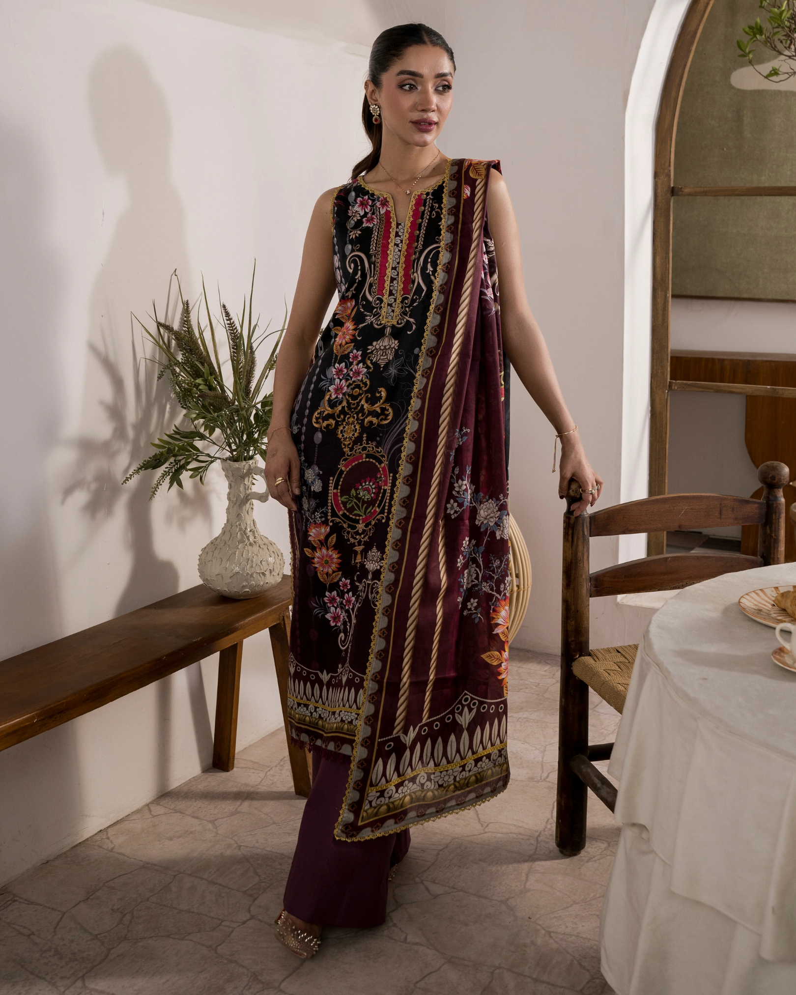 Woman in a traditional embroidered outfit standing in a room with a wooden bench and table.