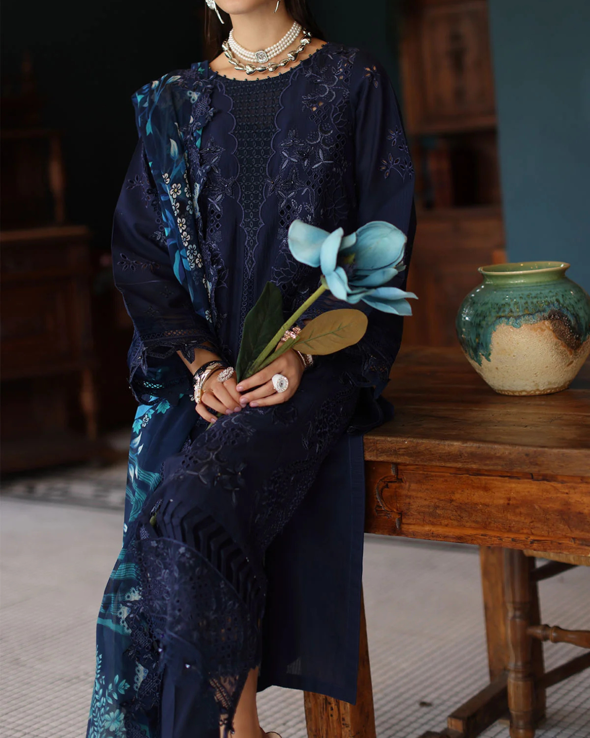 Woman in a dark blue lace dress holding flowers, sitting on a wooden stool.