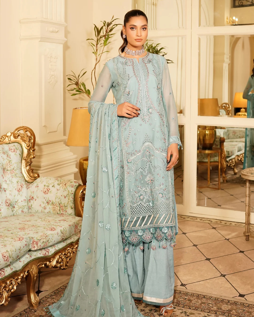 Woman in a light blue embroidered traditional outfit standing in an elegant room.