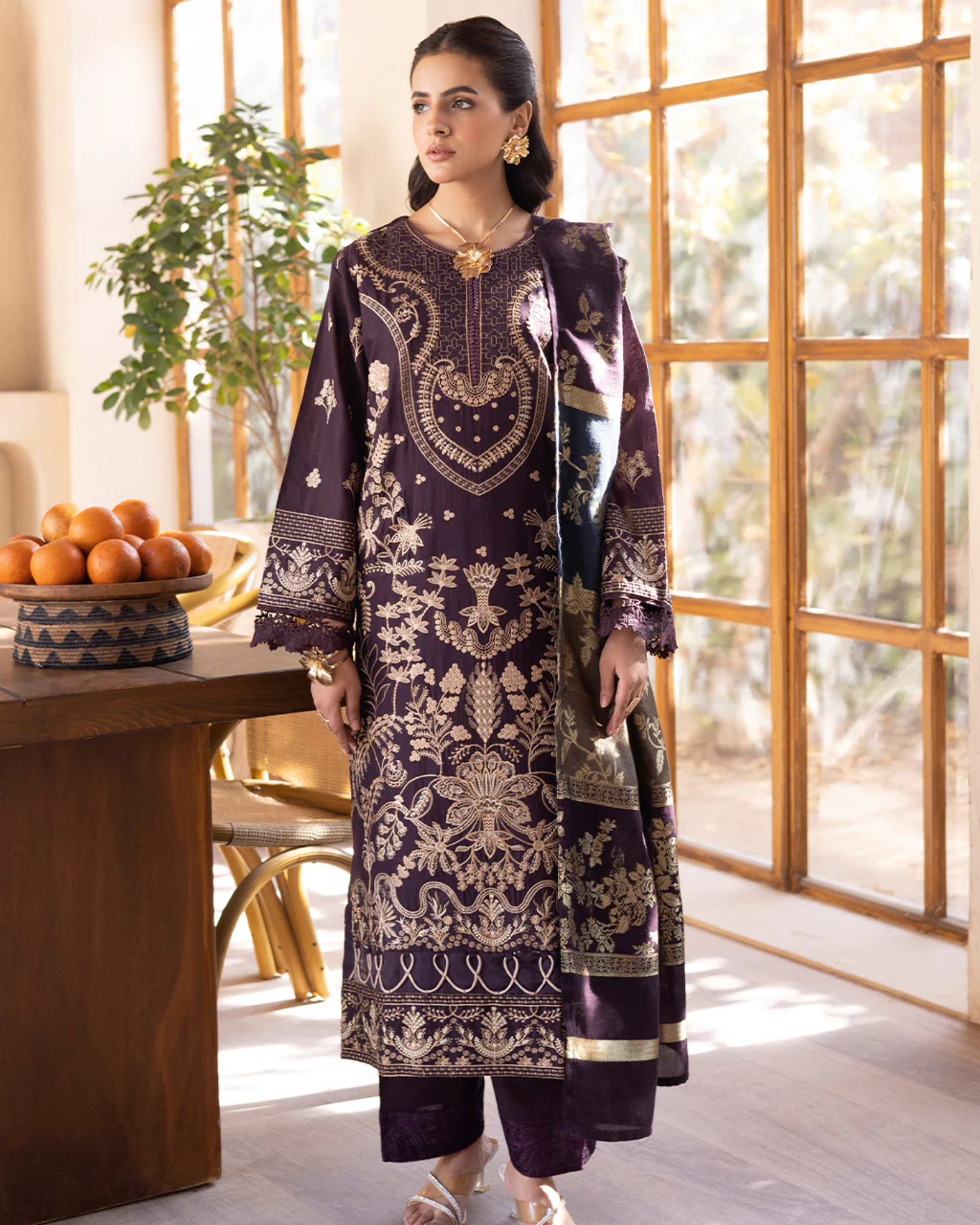 Woman in a dark purple embroidered outfit standing in a bright room with large windows.