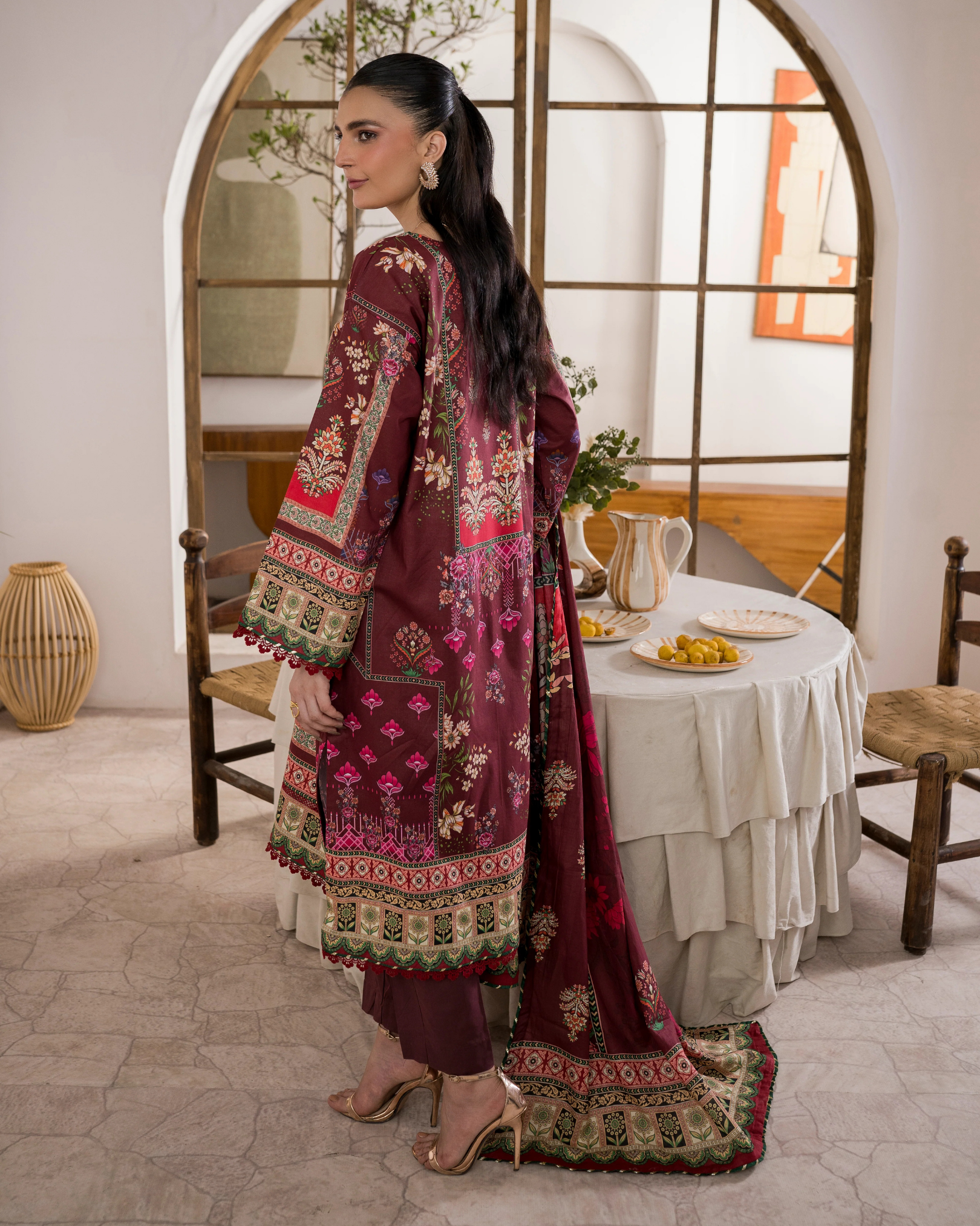 Woman in a burgundy embroidered outfit standing in a room with a table and chairs.