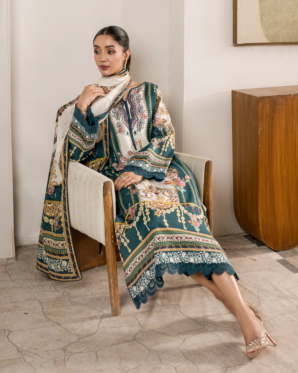 Woman sitting on a chair wearing an ornate, patterned outfit in a room with a wooden table and white walls.
