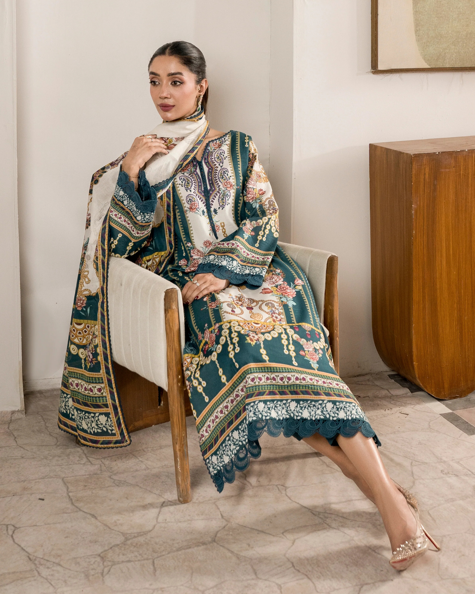 Woman sitting on a chair wearing an ornate, patterned outfit in a room with a wooden table and white walls.