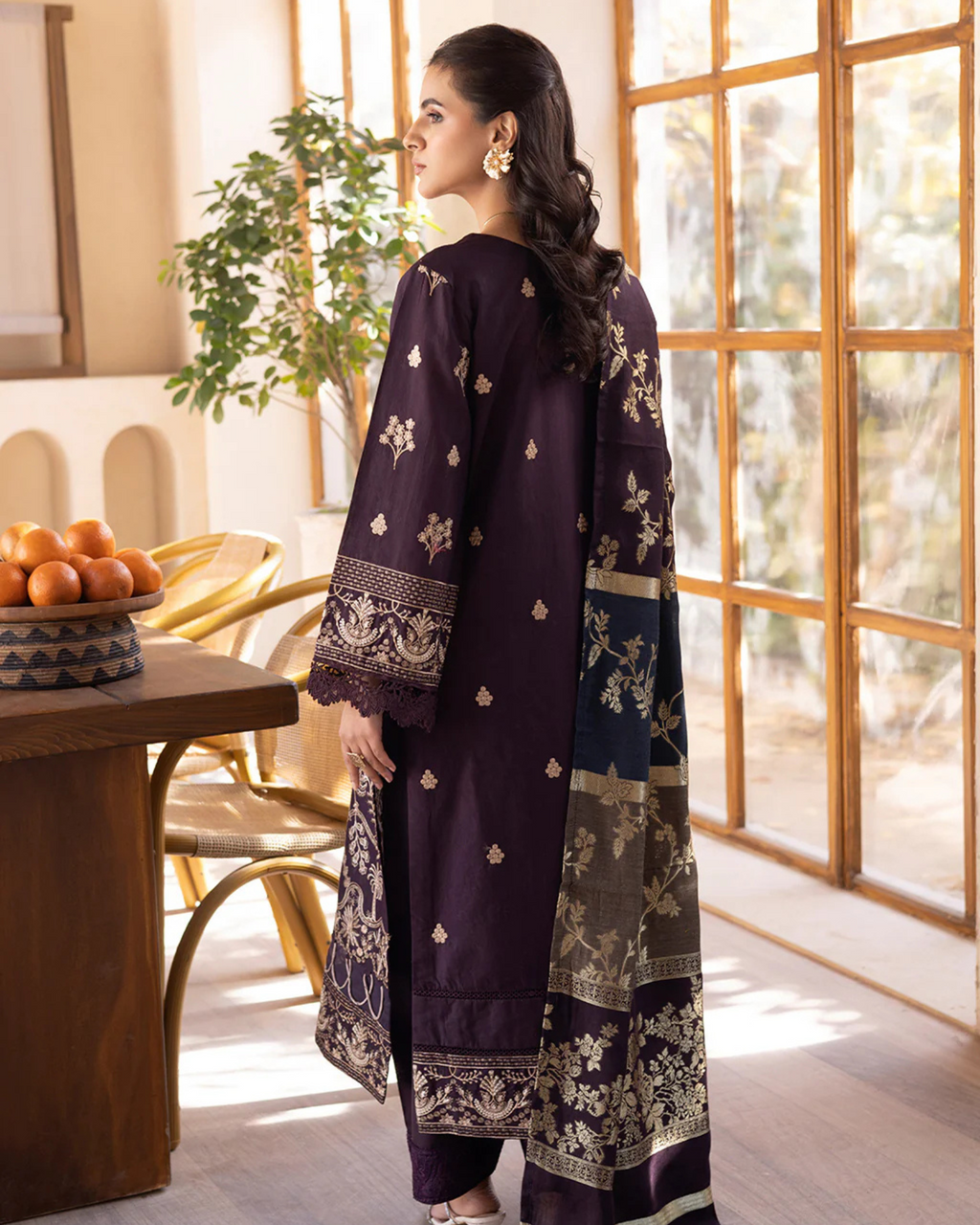 Woman in a dark purple traditional outfit with floral patterns standing in a bright room.
