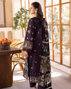 Woman in a dark purple traditional outfit with floral patterns standing in a bright room.