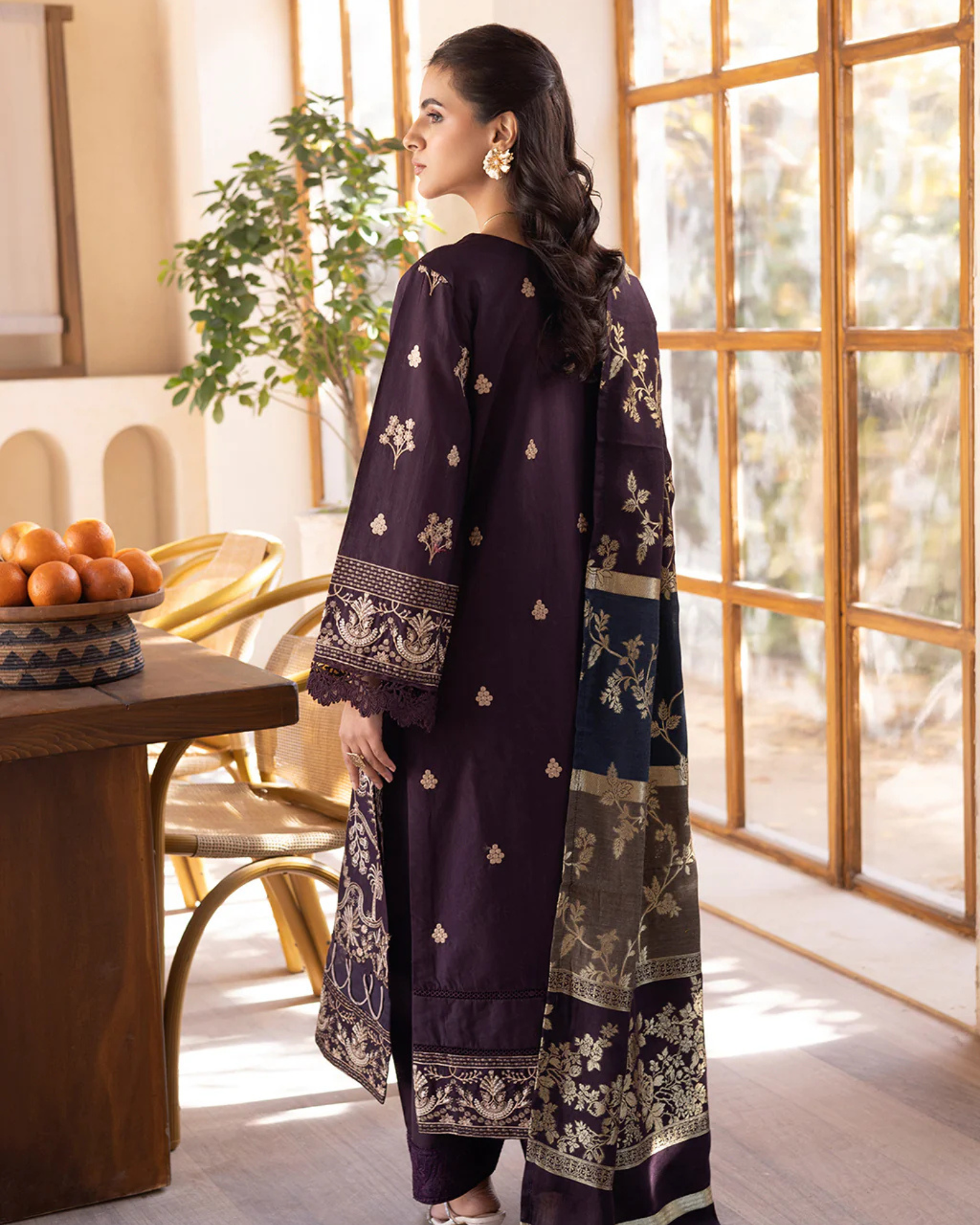 Woman in a dark purple traditional outfit with floral patterns standing in a bright room.