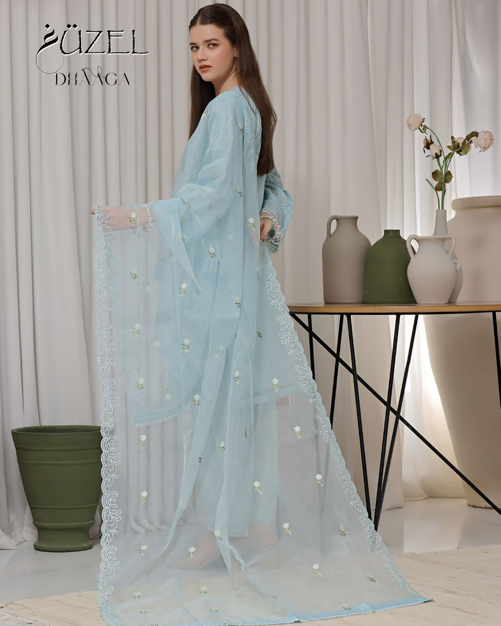 Woman holding a light blue saree with white floral patterns in a minimalistic room.