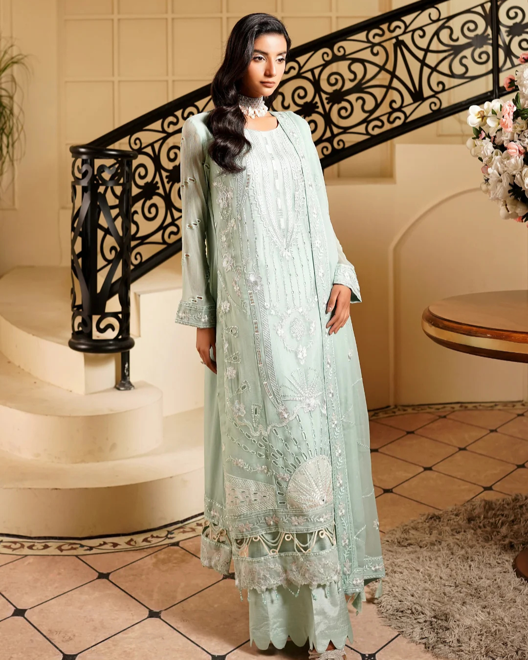 Woman in a light green embroidered dress standing in a decorative interior setting.