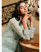 Woman in a light green embroidered outfit sitting on a wooden chair with decorative elements in the background.