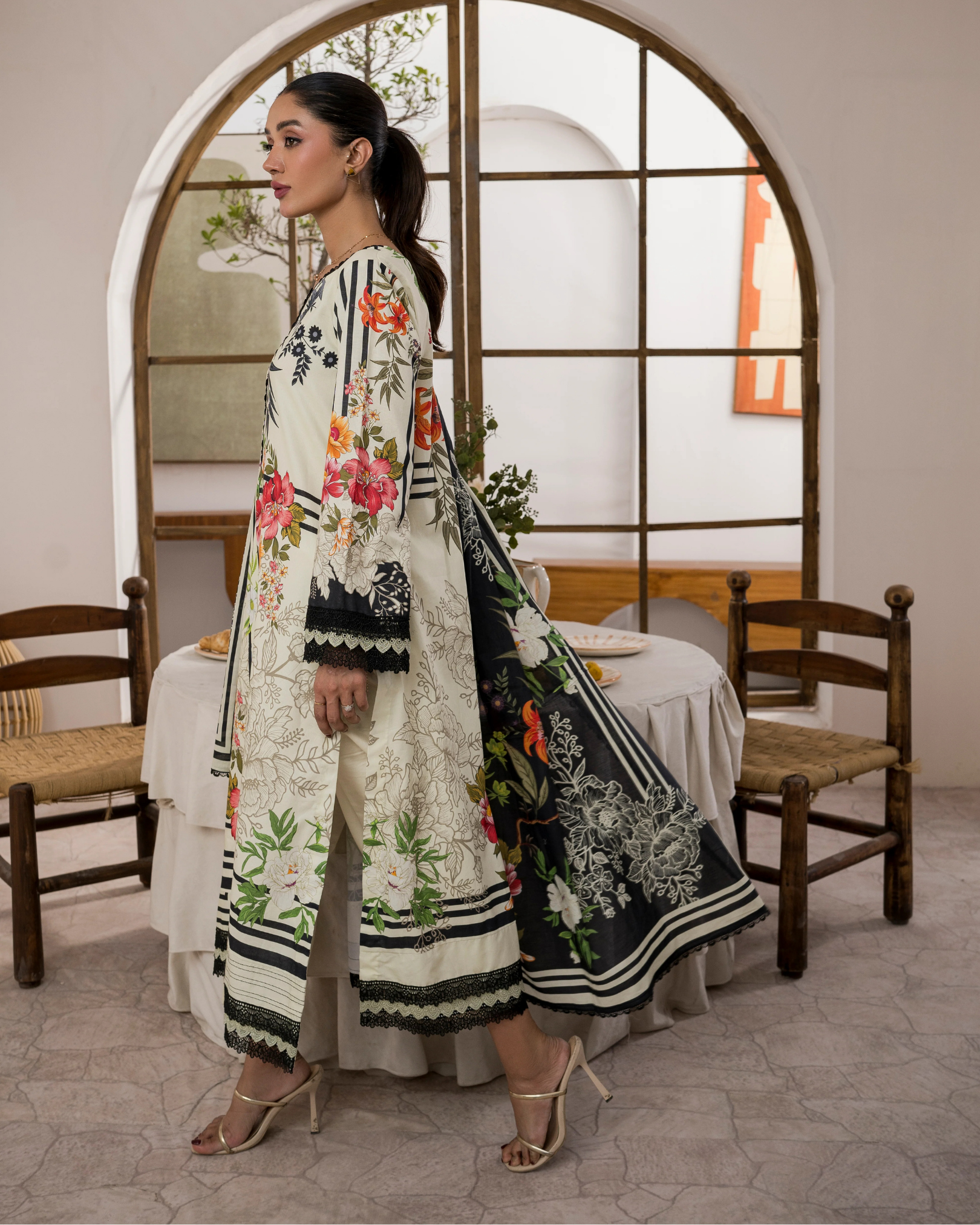 Woman in a floral embroidered outfit standing in a room with wooden chairs and a table.