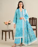 Woman wearing a blue embroidered traditional outfit with white lace details, standing next to a wooden chair and table with flowers.