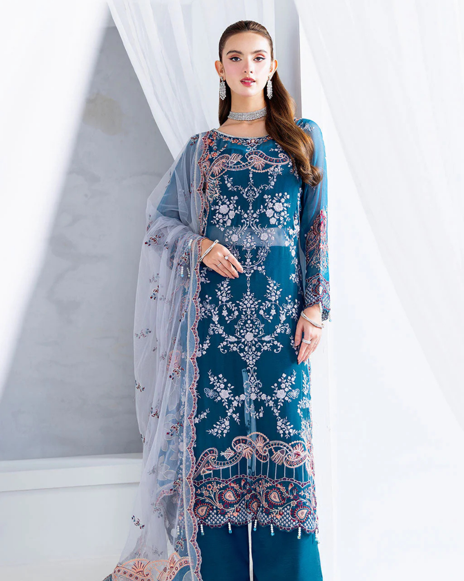 Woman wearing a blue embroidered outfit with a sheer dupatta against a white curtain background