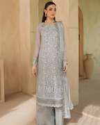 Woman wearing a light gray embroidered traditional outfit in a warm-toned room.