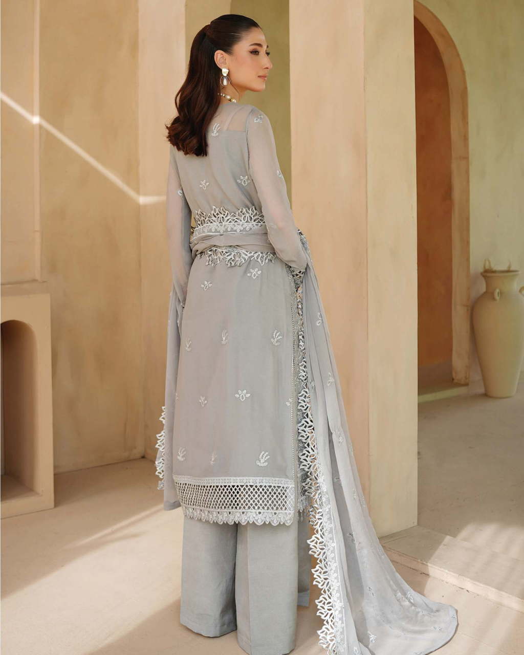 Woman in a light gray traditional outfit with intricate embroidery in a warm-toned room.