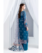 Woman wearing a blue embroidered traditional outfit with a sheer overlay against white curtains.