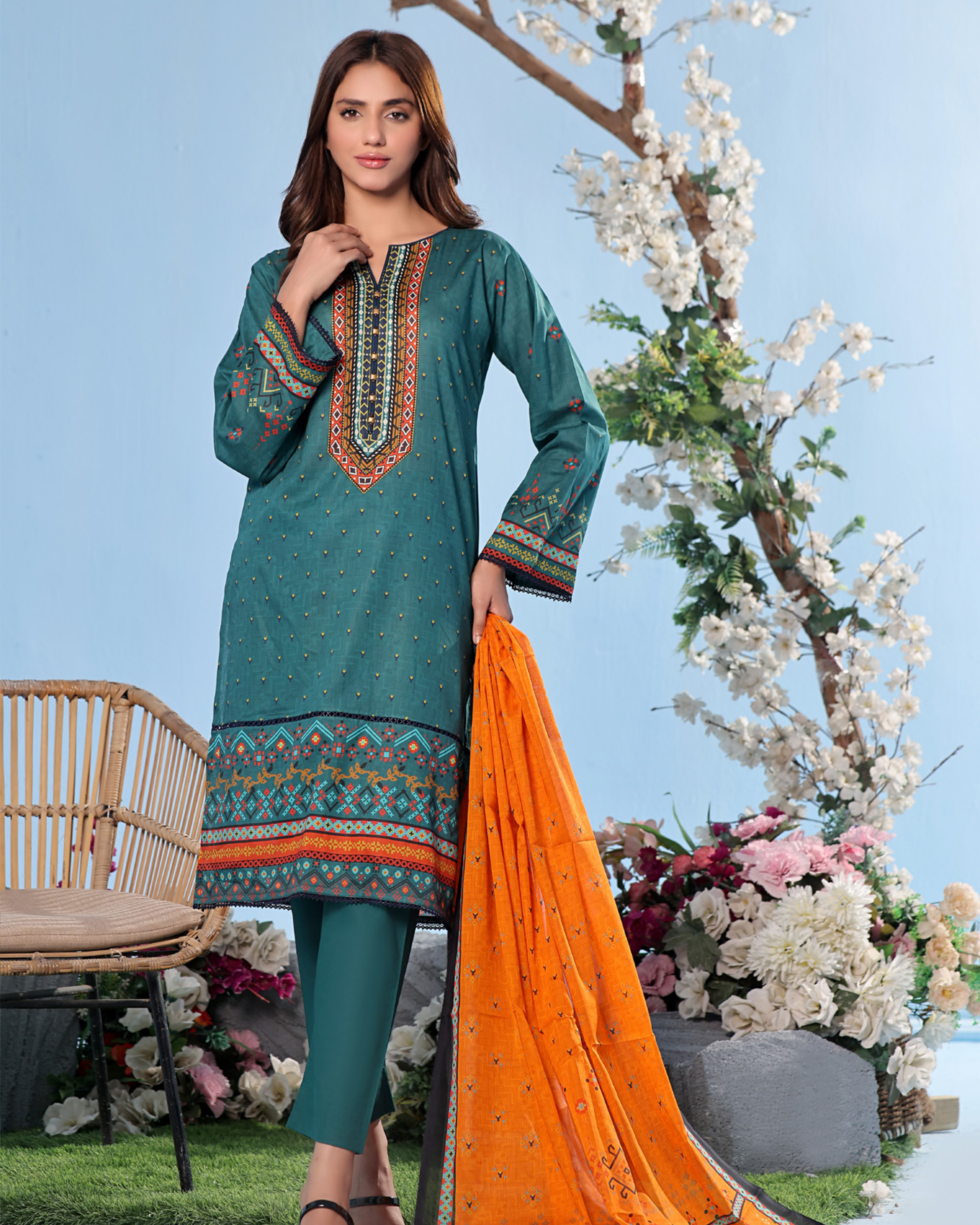 Woman in teal embroidered dress with orange dupatta standing outdoors with flowers and chair in background